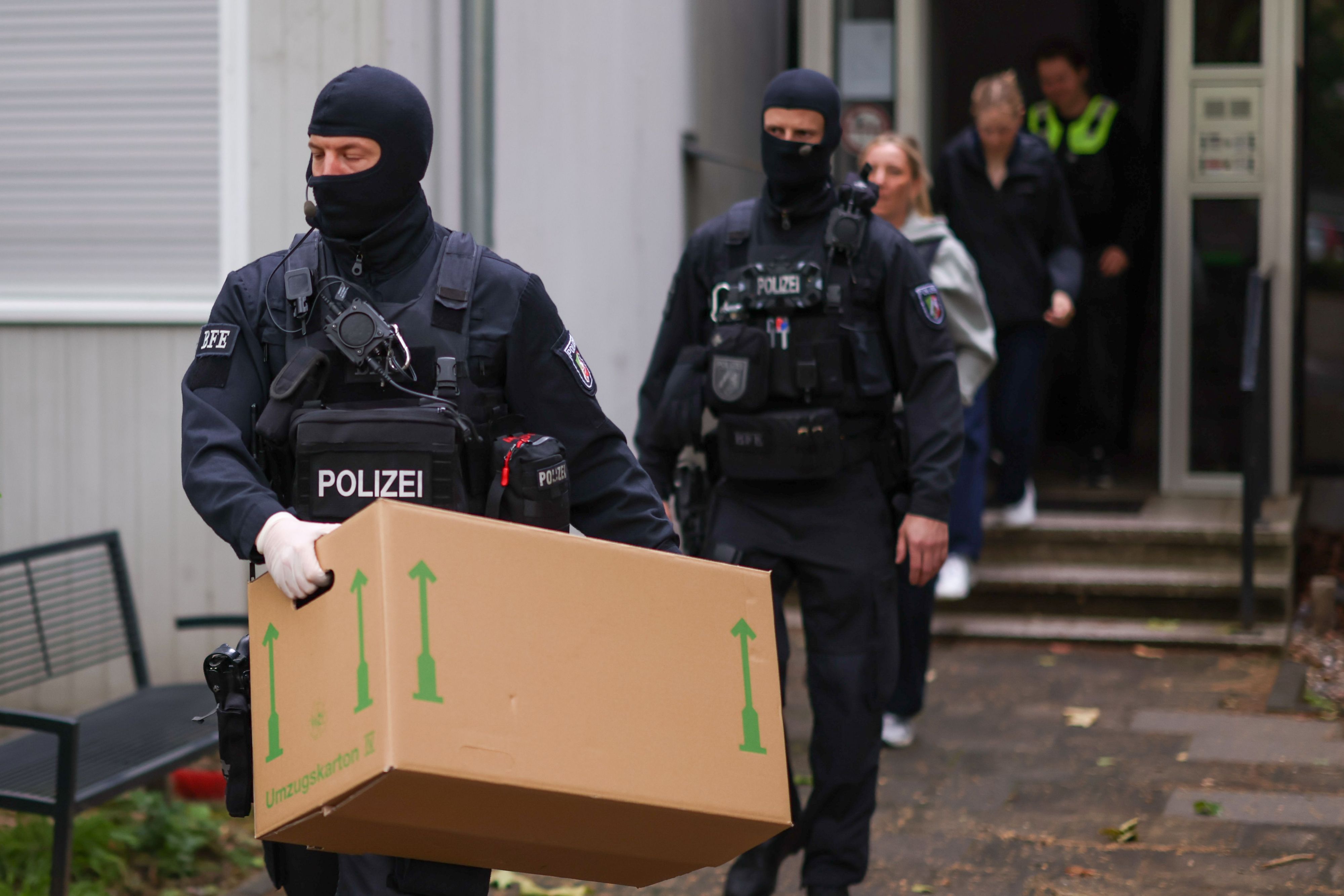 Zwei vermummte Polizisten verlassen ein Gebäude, einer trägt einen großen Karton.
