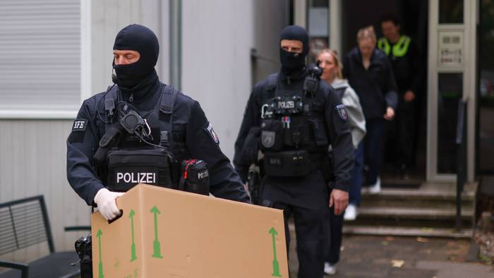 Zwei vermummte Polizisten verlassen ein Gebäude, einer trägt einen großen Karton.