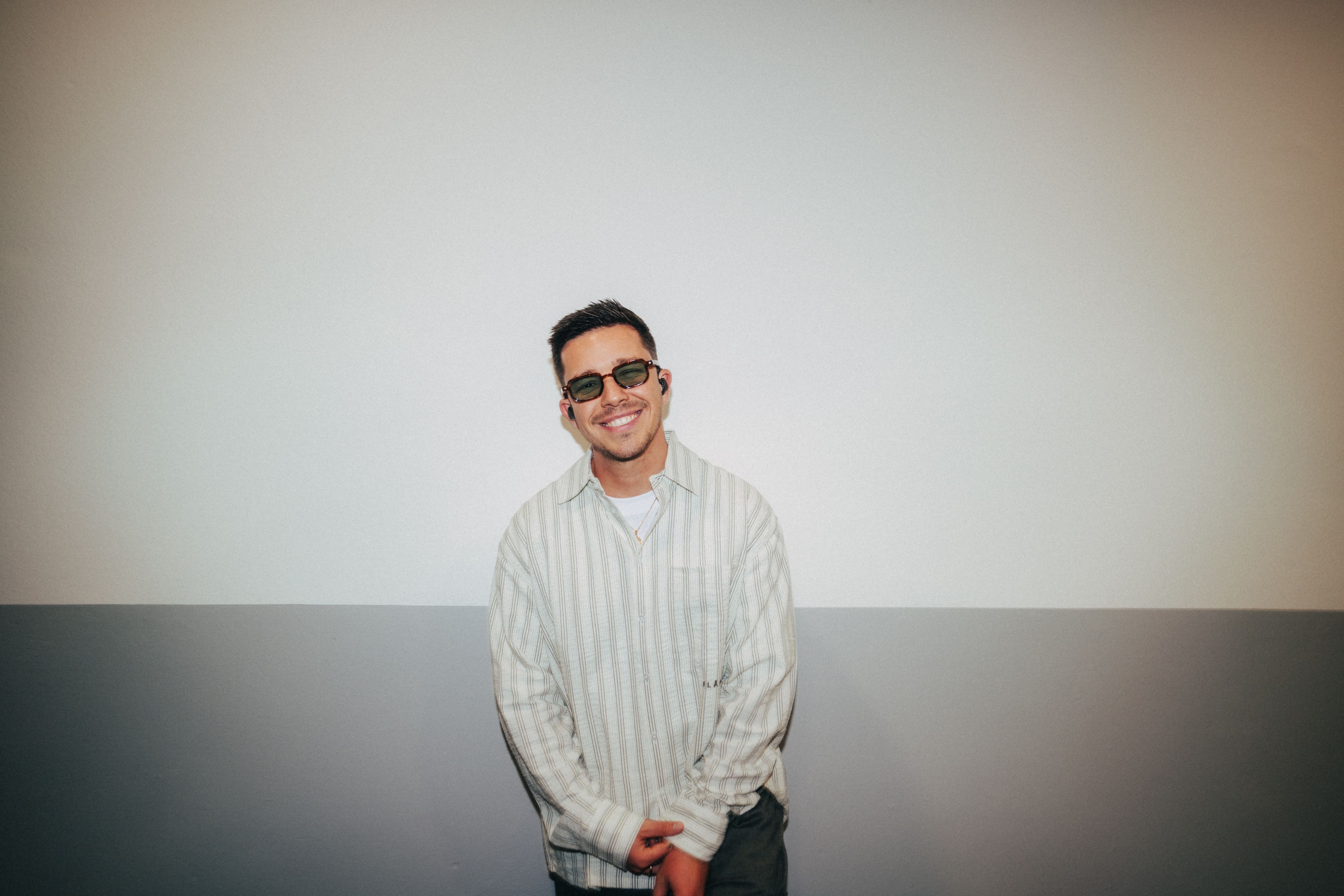 Nico Santos mit dunkler Brille und In-Ears lacht in die Kamera. Er trägt ein gestreiftes Hemd.
