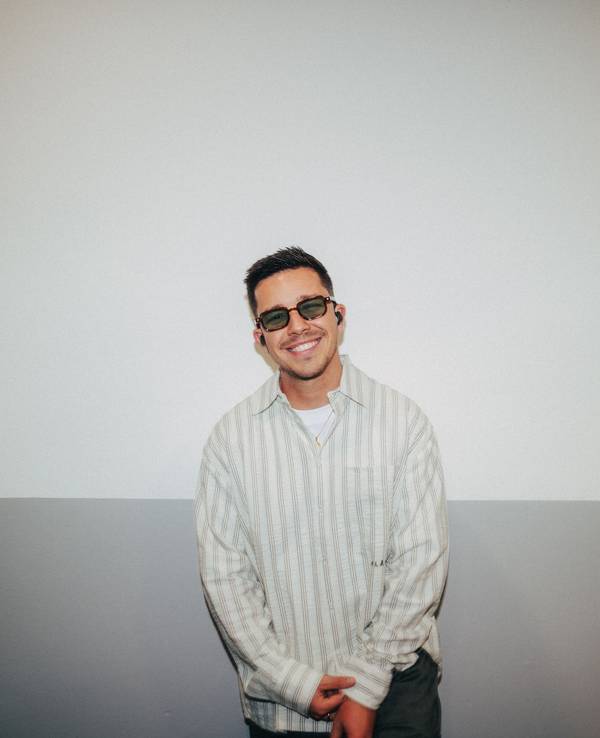 Nico Santos mit dunkler Brille und In-Ears lacht in die Kamera. Er trägt ein gestreiftes Hemd.