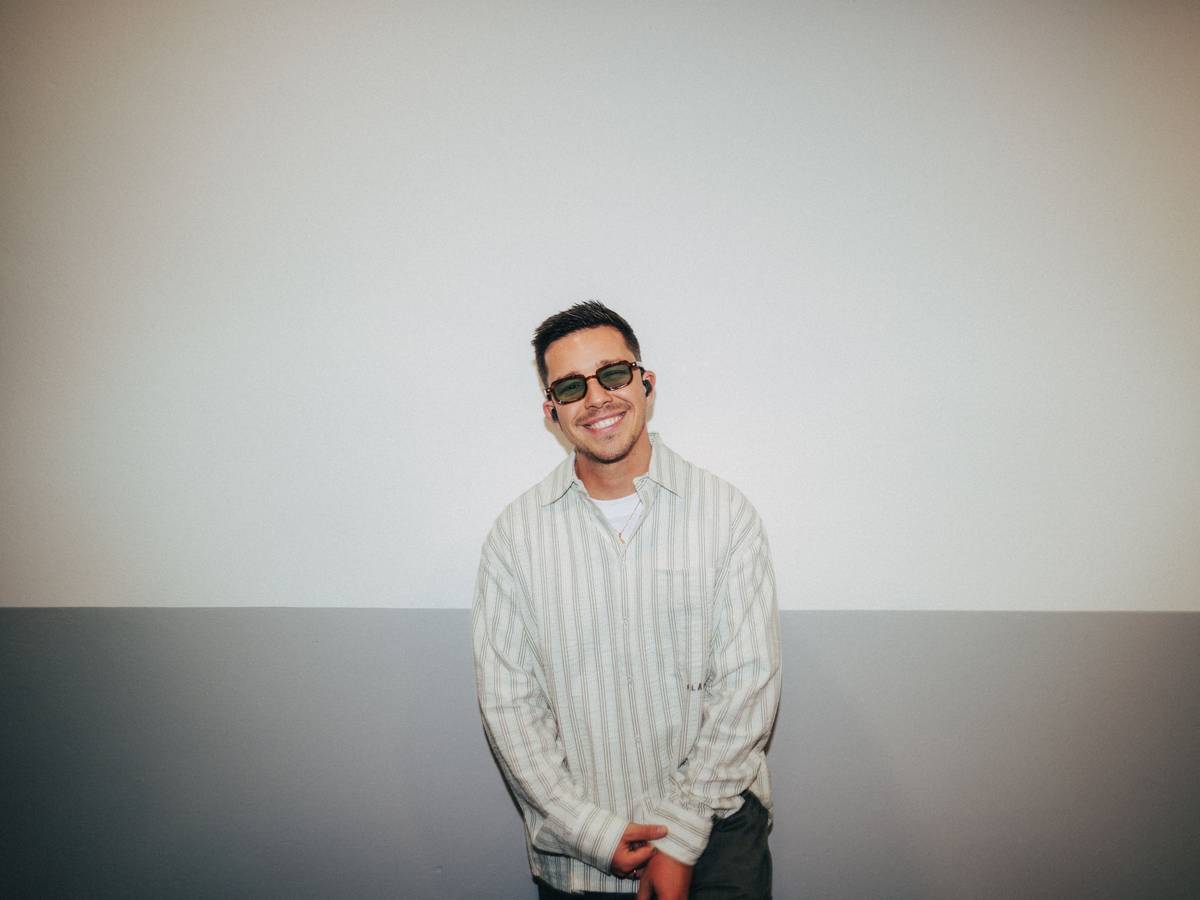 Nico Santos mit dunkler Brille und In-Ears lacht in die Kamera. Er trägt ein gestreiftes Hemd.