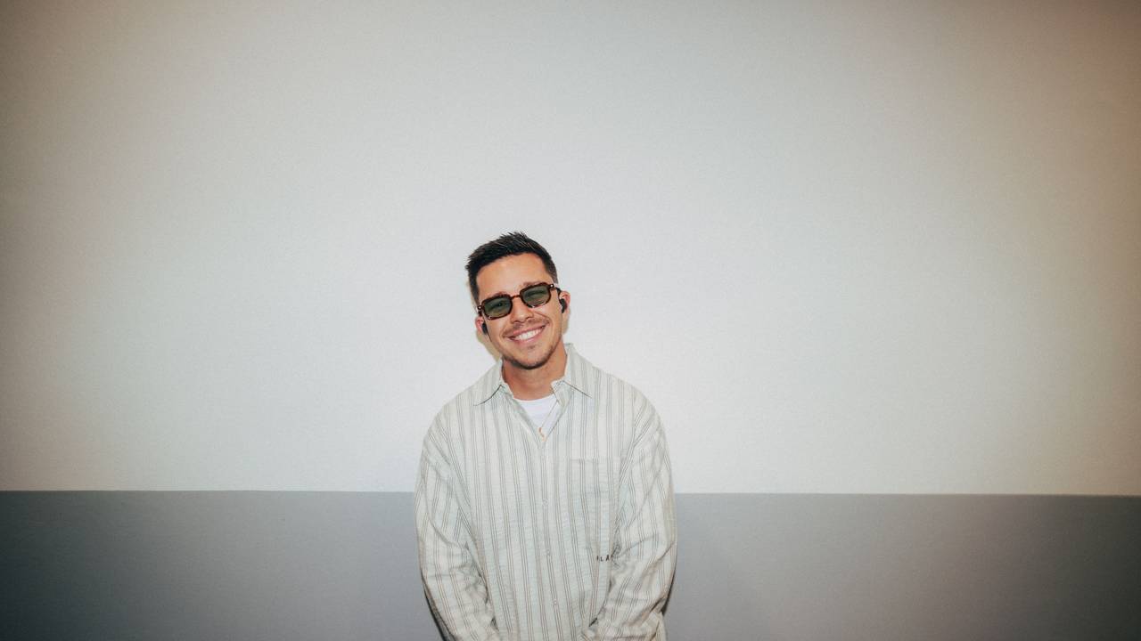 Nico Santos mit dunkler Brille und In-Ears lacht in die Kamera. Er trägt ein gestreiftes Hemd.
