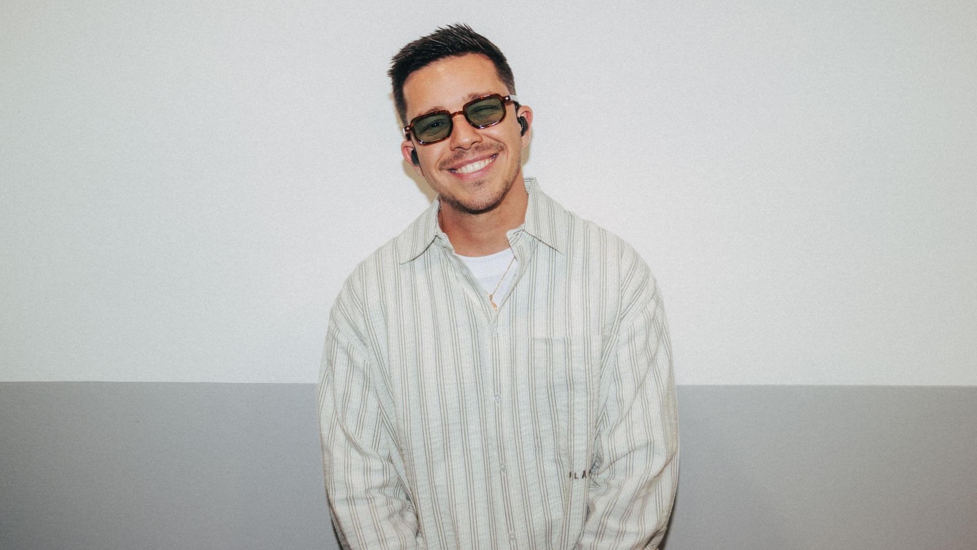 Nico Santos mit dunkler Brille und In-Ears lacht in die Kamera. Er trägt ein gestreiftes Hemd.