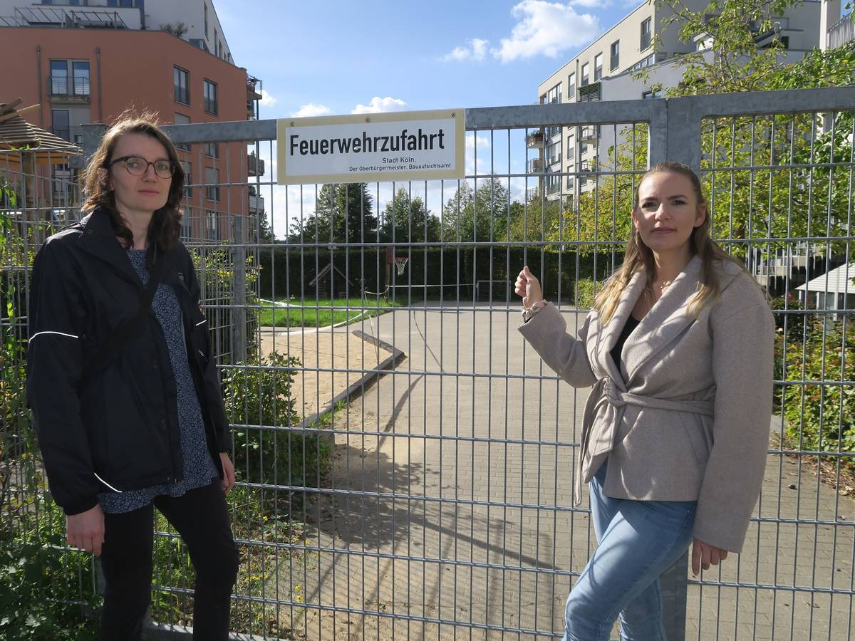 Zwei Frauen stehen vor einem abgesperrten Spielplatz; eine zeigt auf das Tor mit dem Schild „Feuerwehrzufahrt“.