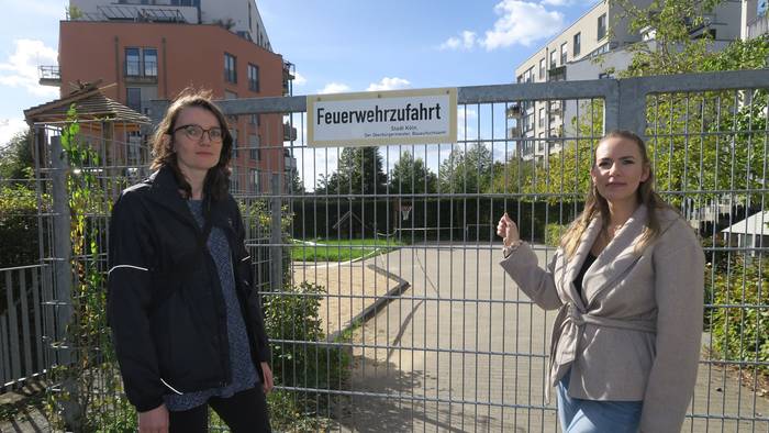 Zwei Frauen stehen vor einem abgesperrten Spielplatz; eine zeigt auf das Tor mit dem Schild „Feuerwehrzufahrt“.