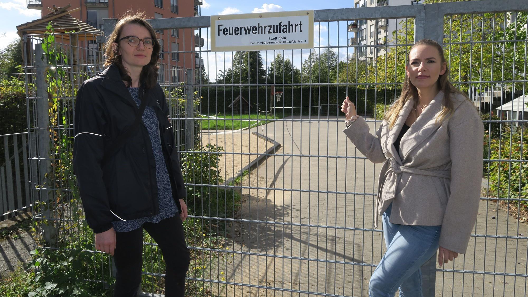 Zwei Frauen stehen vor einem abgesperrten Spielplatz; eine zeigt auf das Tor mit dem Schild „Feuerwehrzufahrt“.