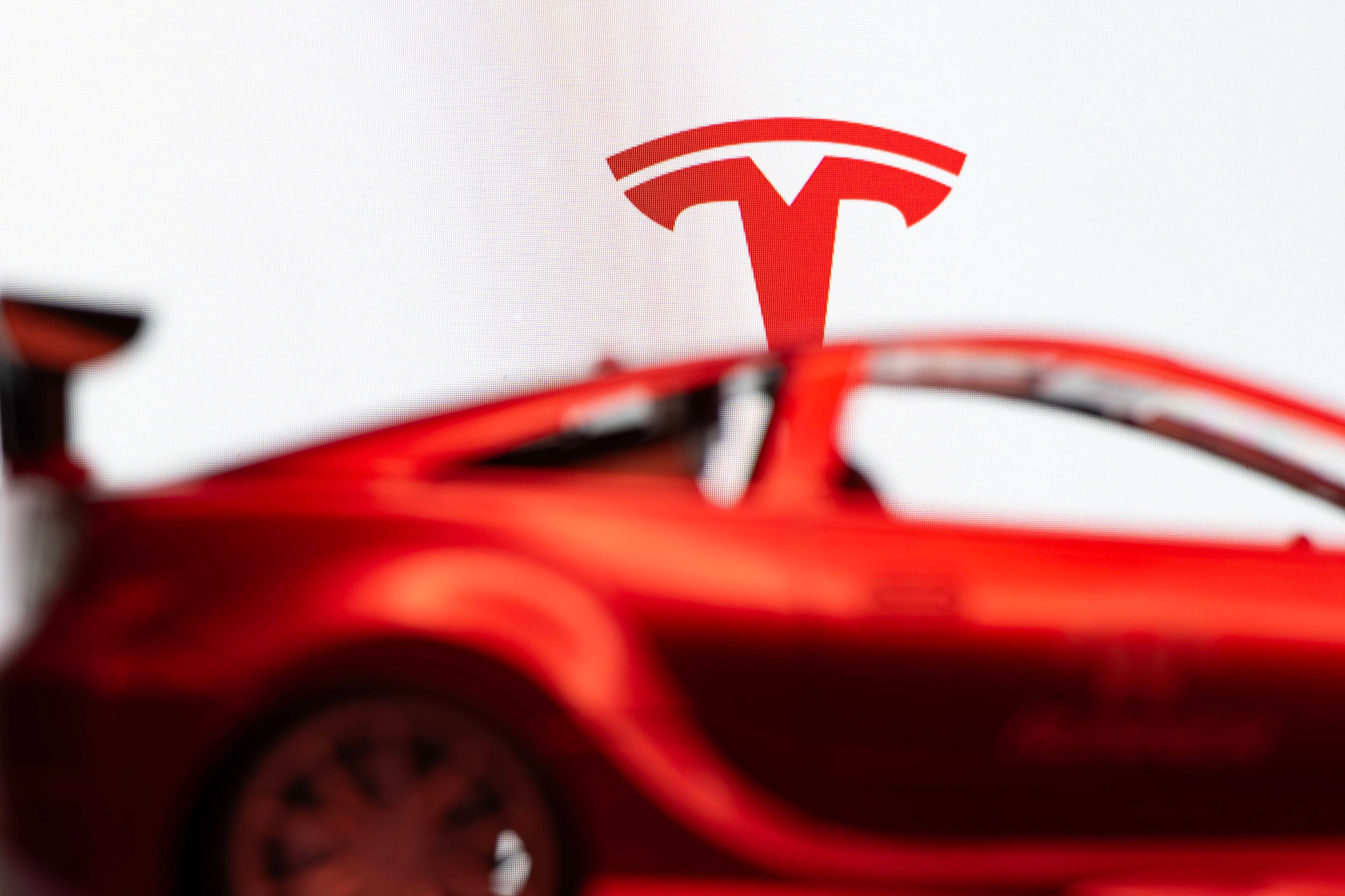 Das Tesla-T-Symbol über einem verschwommenen roten Auto vor weißem Hintergrund.