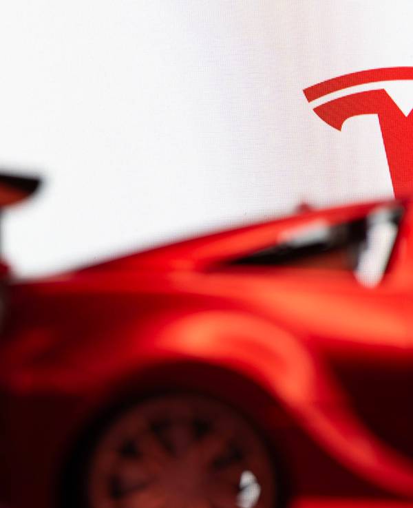 Das Tesla-T-Symbol über einem verschwommenen roten Auto vor weißem Hintergrund.
