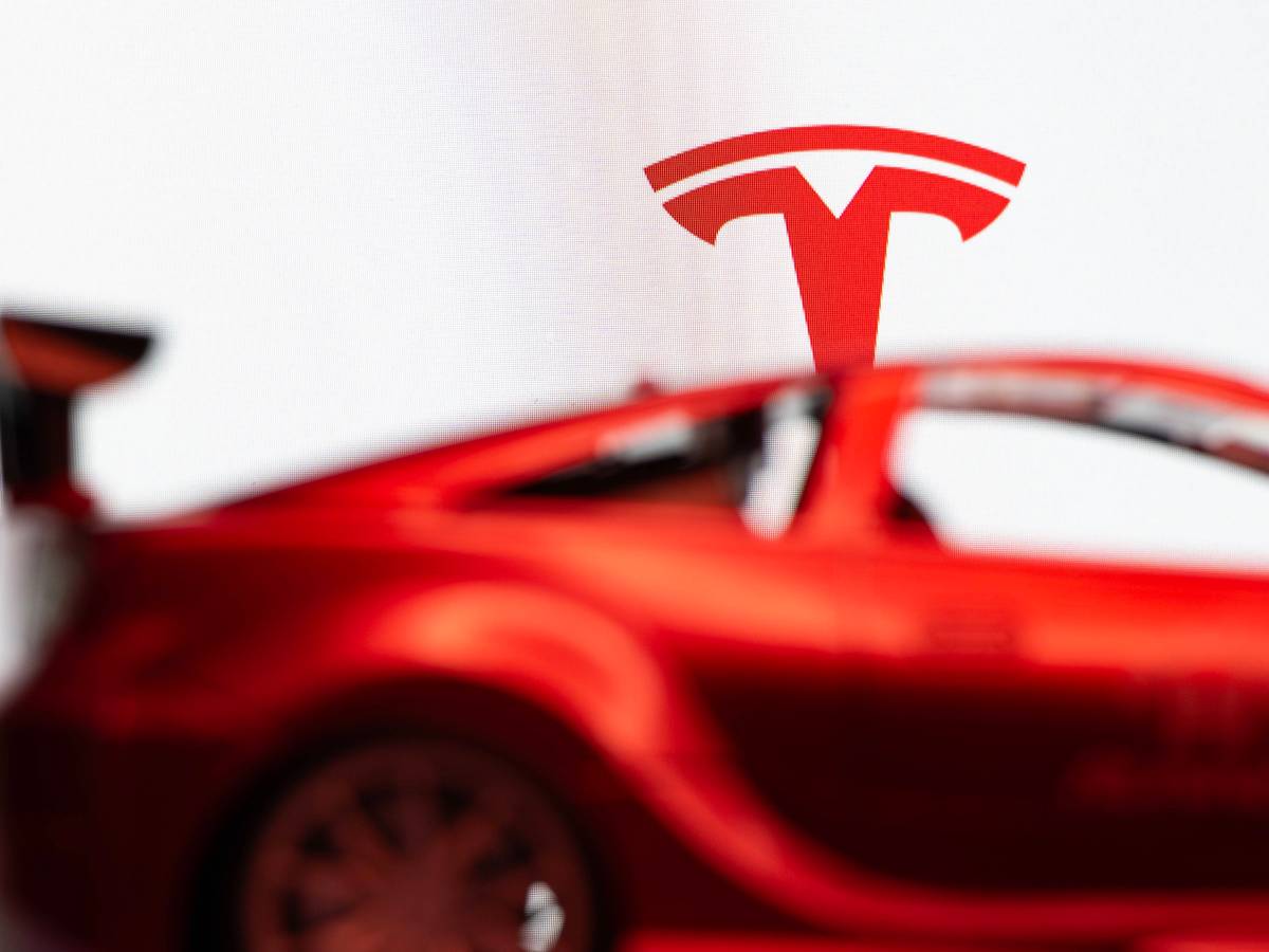 Das Tesla-T-Symbol über einem verschwommenen roten Auto vor weißem Hintergrund.