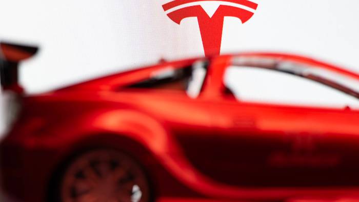 Das Tesla-T-Symbol über einem verschwommenen roten Auto vor weißem Hintergrund.
