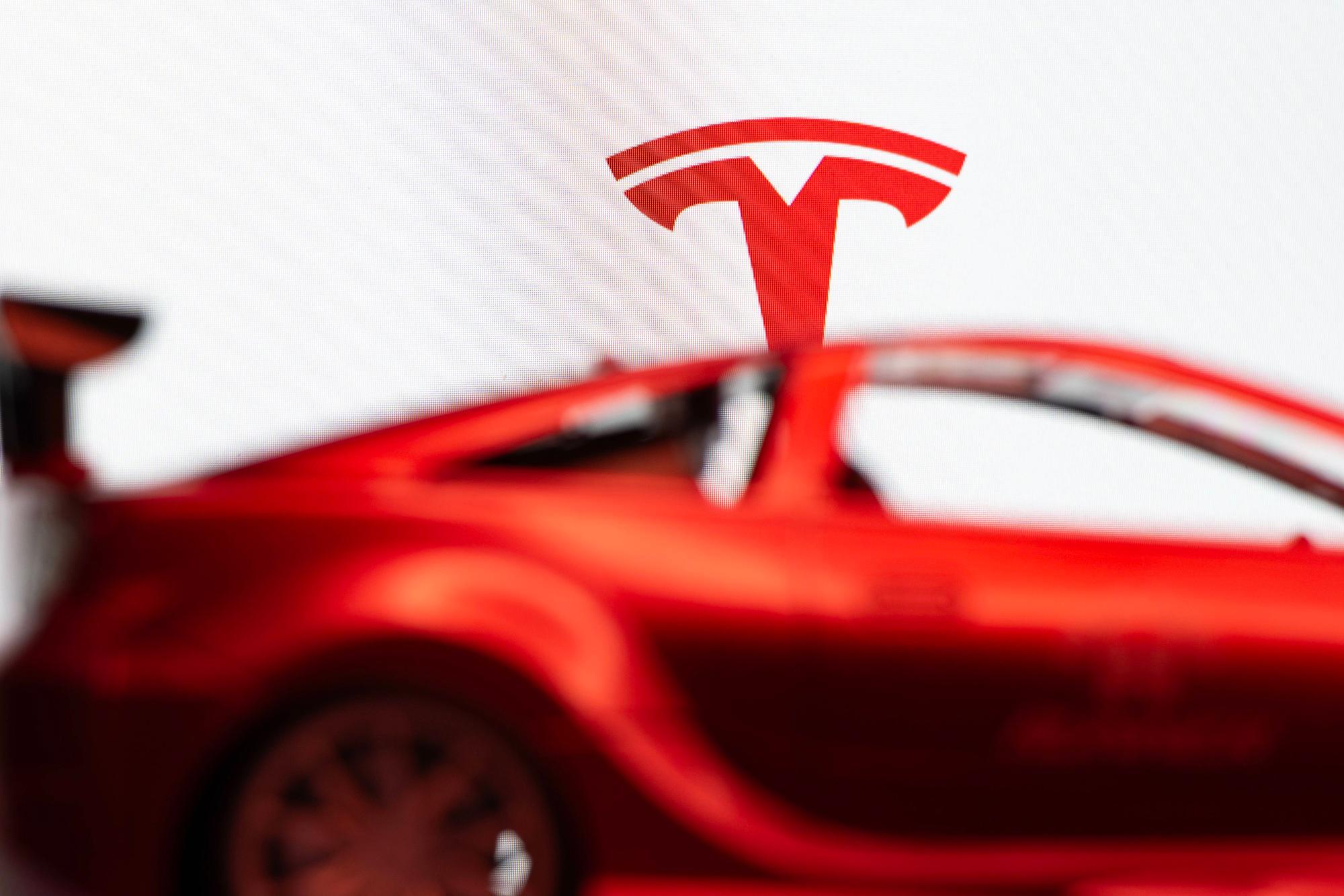 Das Tesla-T-Symbol über einem verschwommenen roten Auto vor weißem Hintergrund.