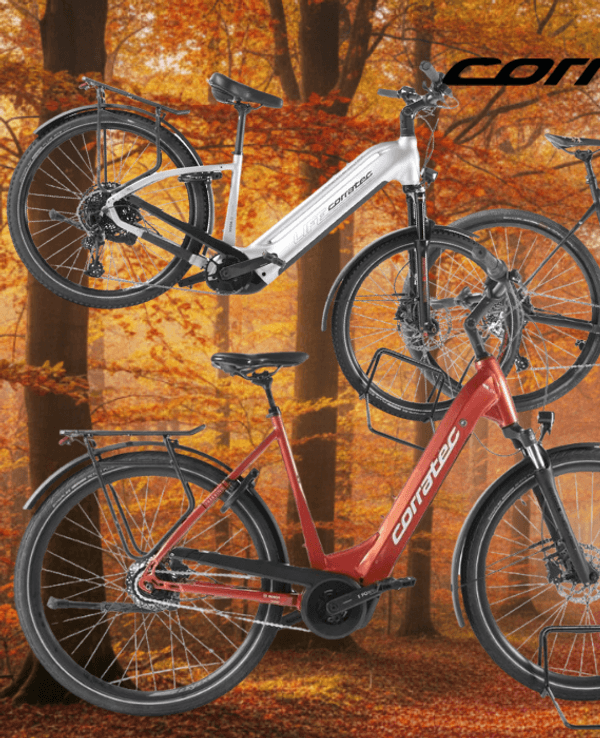 Mehrere Corratec E‑Bikes unterschiedlicher Typen – darunter Trekking-, City- und Mountainbikes – vor einer Landstraße an einem sonnigen Tag in Natur.