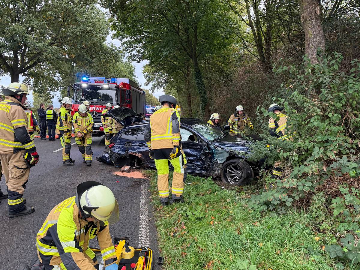 Einsatzkräfte der Feuerwehr sind an einem stark deformierten Auto im Einsatz. Der Wagen war infolge eines Unfalls seitlich gegen einen Baum geprallt.