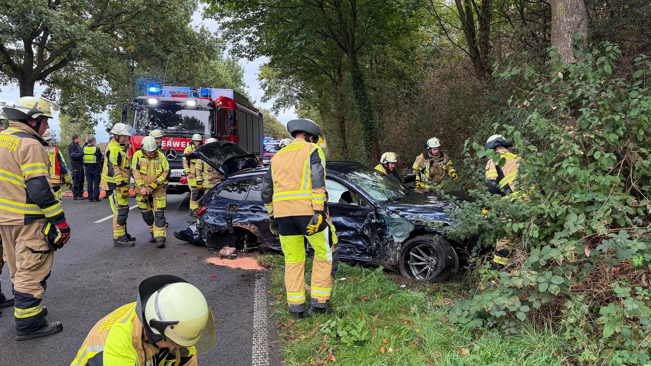 Einsatzkräfte der Feuerwehr sind an einem stark deformierten Auto im Einsatz. Der Wagen war infolge eines Unfalls seitlich gegen einen Baum geprallt.