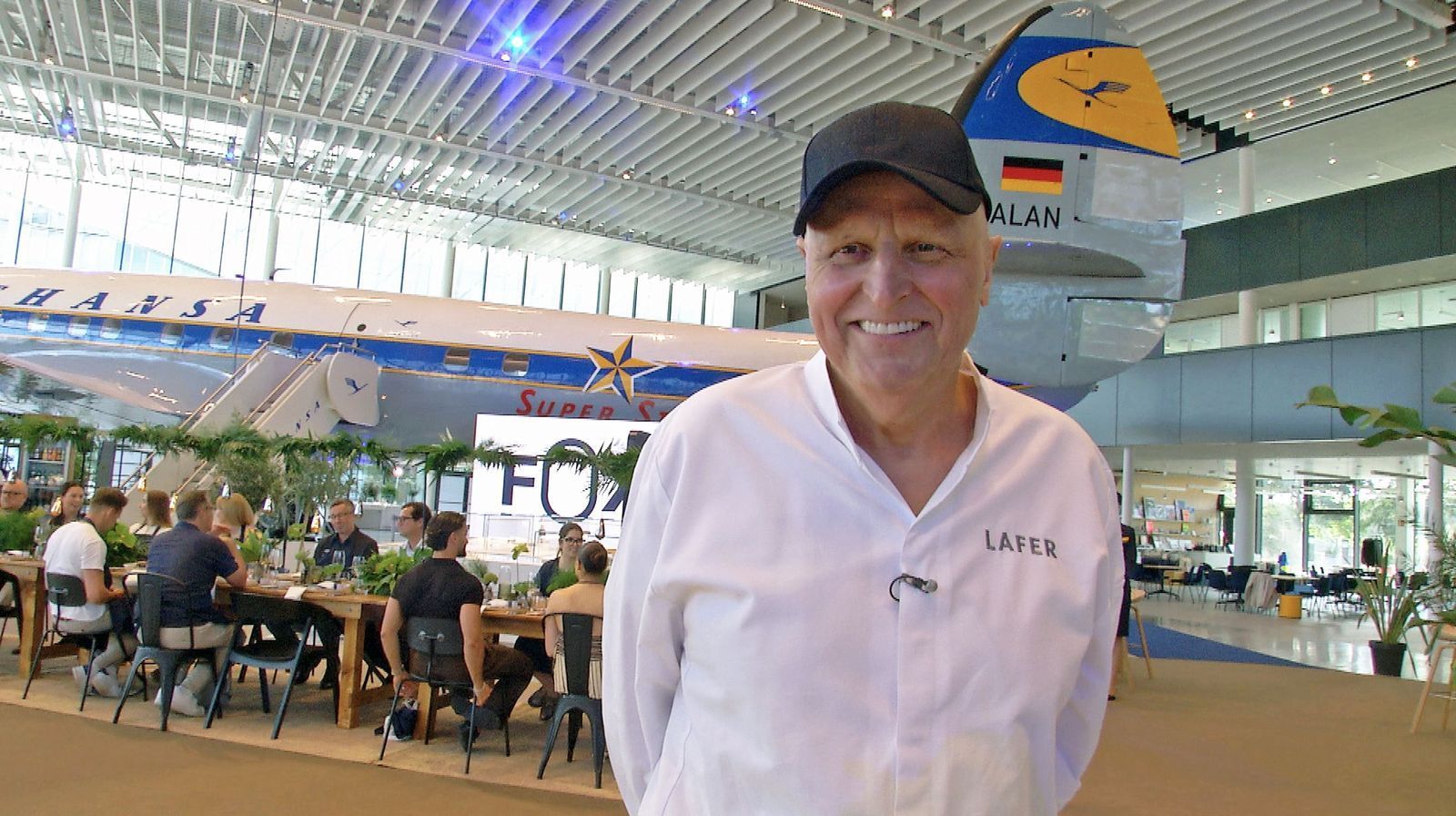 Der markante Schnäuzer fiel zwischenzeitlich der Chemotherapie zum Opfer: Johann Lafer im Hangar One in der der HR-Sendung „Mittendrin Flughafen Frankfurt“ (abrufbar in der Mediathek). (Bild: HR)