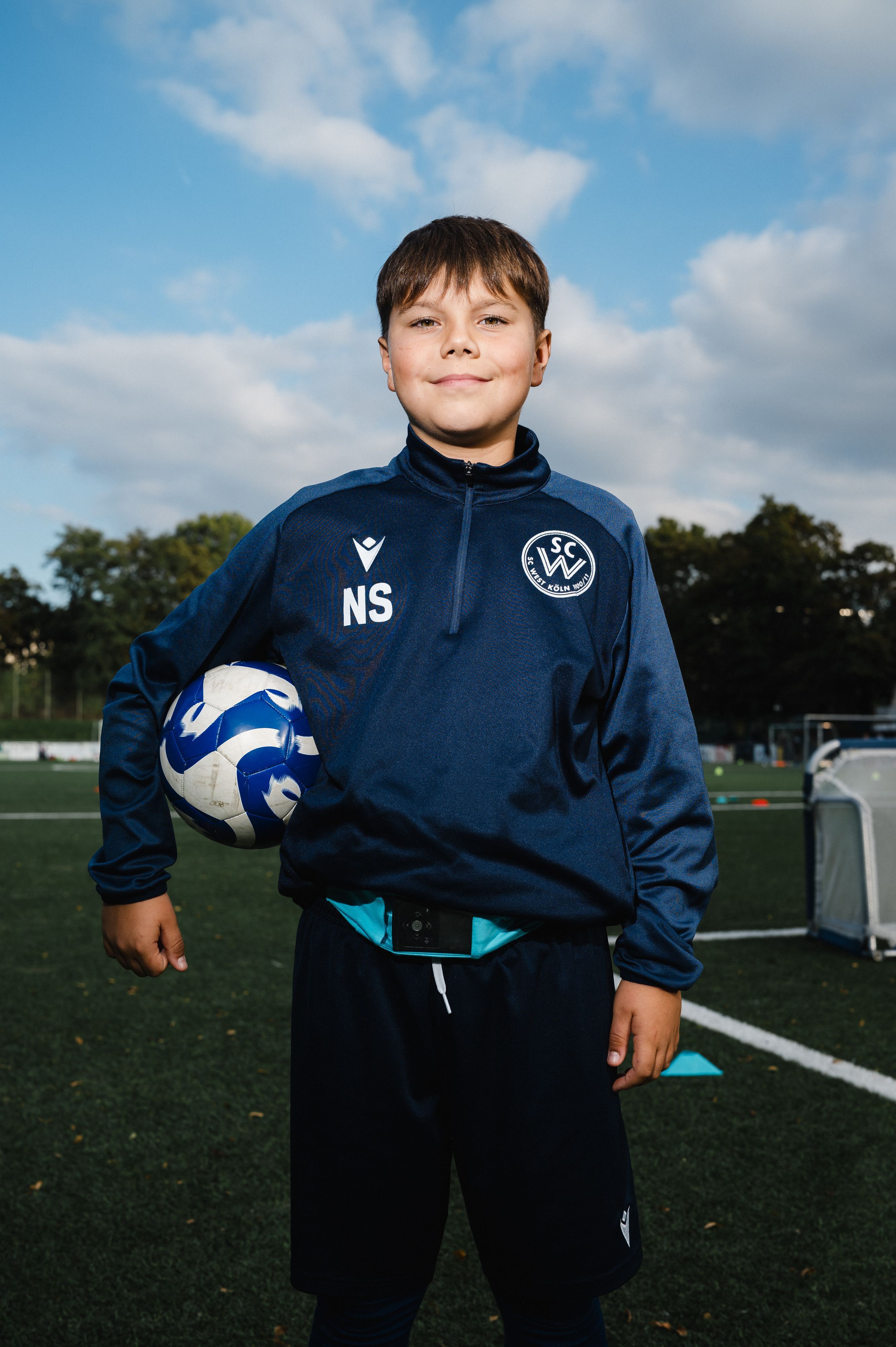 Nick (11) steht auf dem Fußballplatz und hat sich einen Fußball unter den rechten Arm geklemmt.