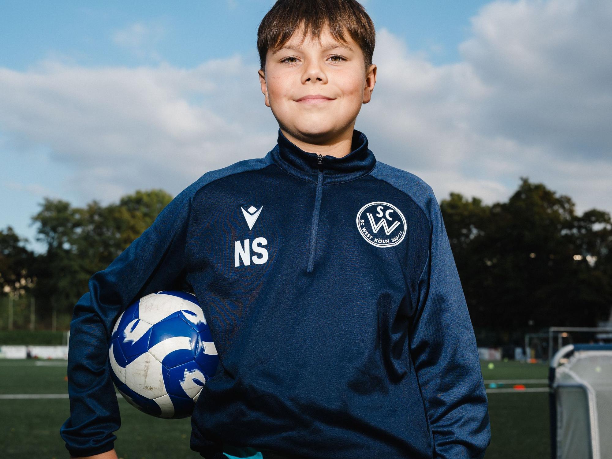 Nick (11) steht auf dem Fußballplatz und hat sich einen Fußball unter den rechten Arm geklemmt.