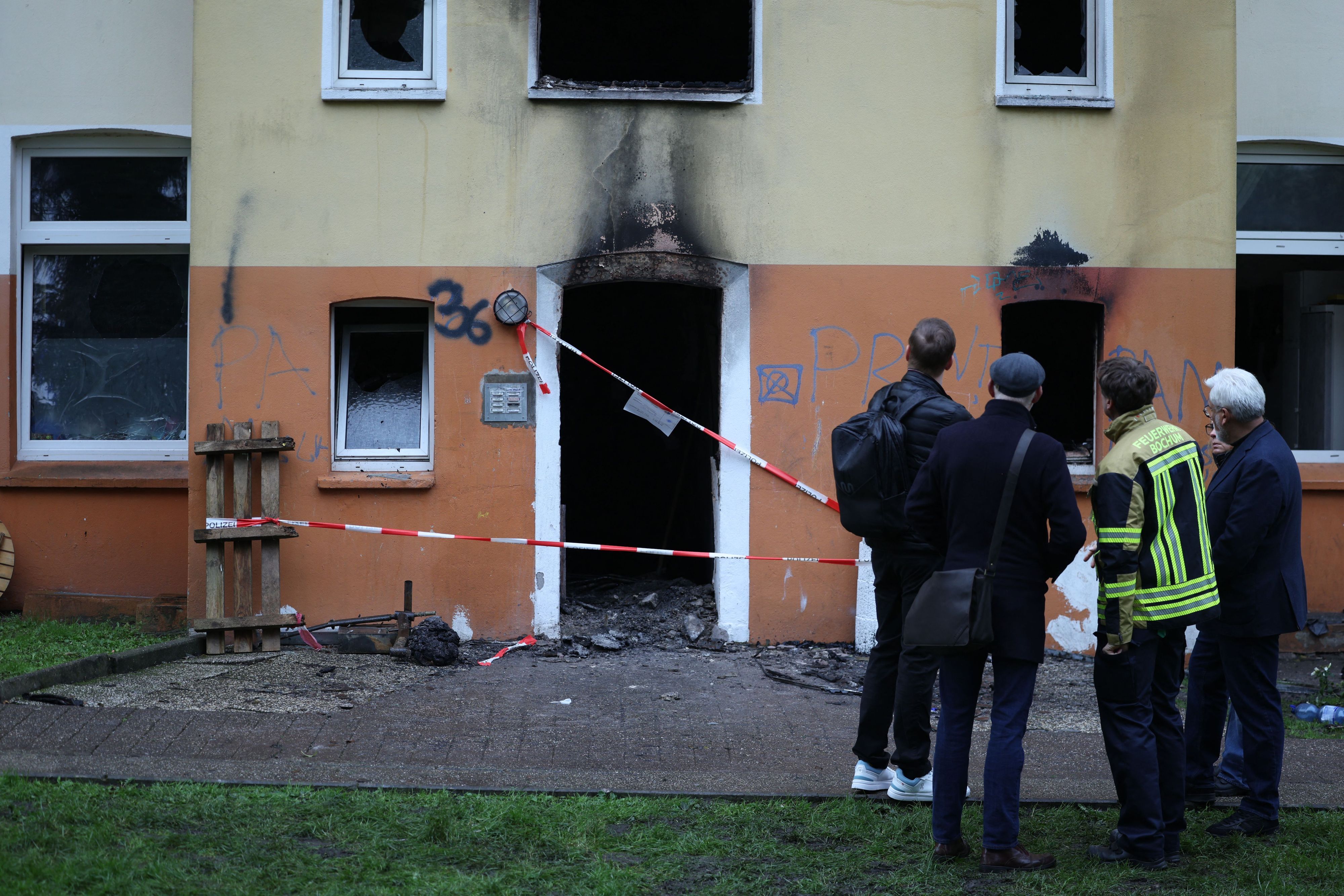 Vier Männer, einer in Feuerwehrjacke, stehe vor dem Mehrfamilienhaus, in dem es gebrannt hat. Der Eingang ist mit Polizei-Flatterband abgesperrt, an der Fassade klebt Ruß.