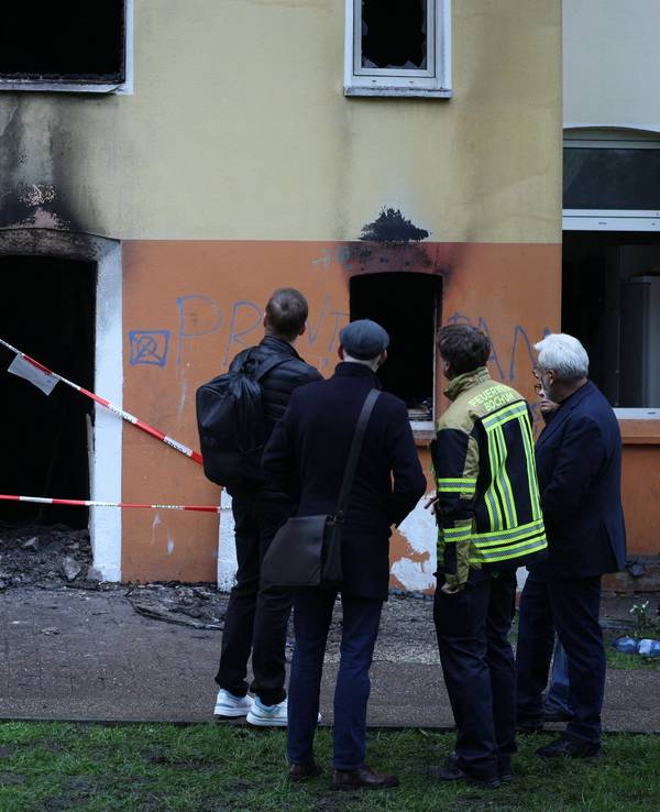 Vier Männer, einer in Feuerwehrjacke, stehe vor dem Mehrfamilienhaus, in dem es gebrannt hat. Der Eingang ist mit Polizei-Flatterband abgesperrt, an der Fassade klebt Ruß.