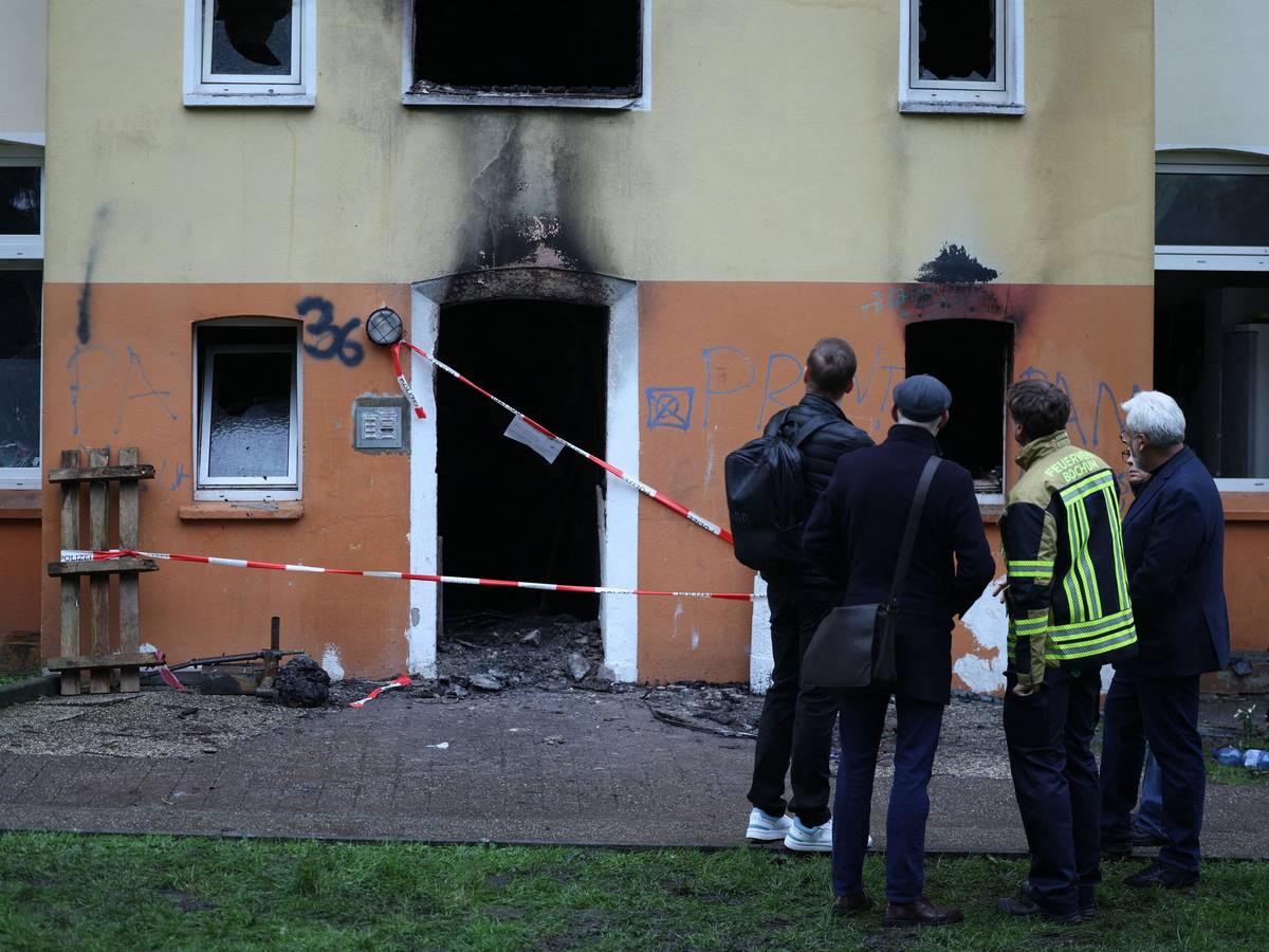 Vier Männer, einer in Feuerwehrjacke, stehe vor dem Mehrfamilienhaus, in dem es gebrannt hat. Der Eingang ist mit Polizei-Flatterband abgesperrt, an der Fassade klebt Ruß.