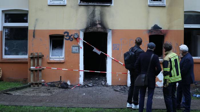 Vier Männer, einer in Feuerwehrjacke, stehe vor dem Mehrfamilienhaus, in dem es gebrannt hat. Der Eingang ist mit Polizei-Flatterband abgesperrt, an der Fassade klebt Ruß.