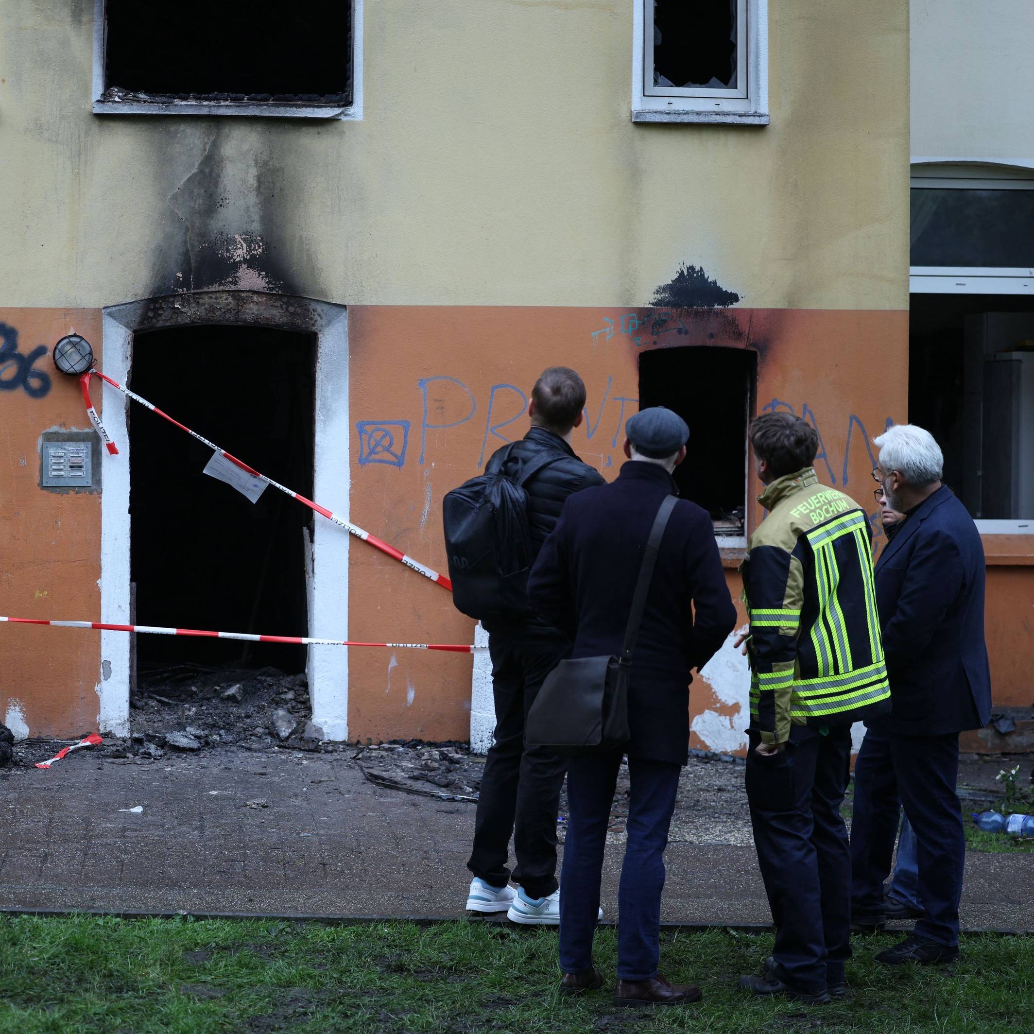 Vier Männer, einer in Feuerwehrjacke, stehe vor dem Mehrfamilienhaus, in dem es gebrannt hat. Der Eingang ist mit Polizei-Flatterband abgesperrt, an der Fassade klebt Ruß.