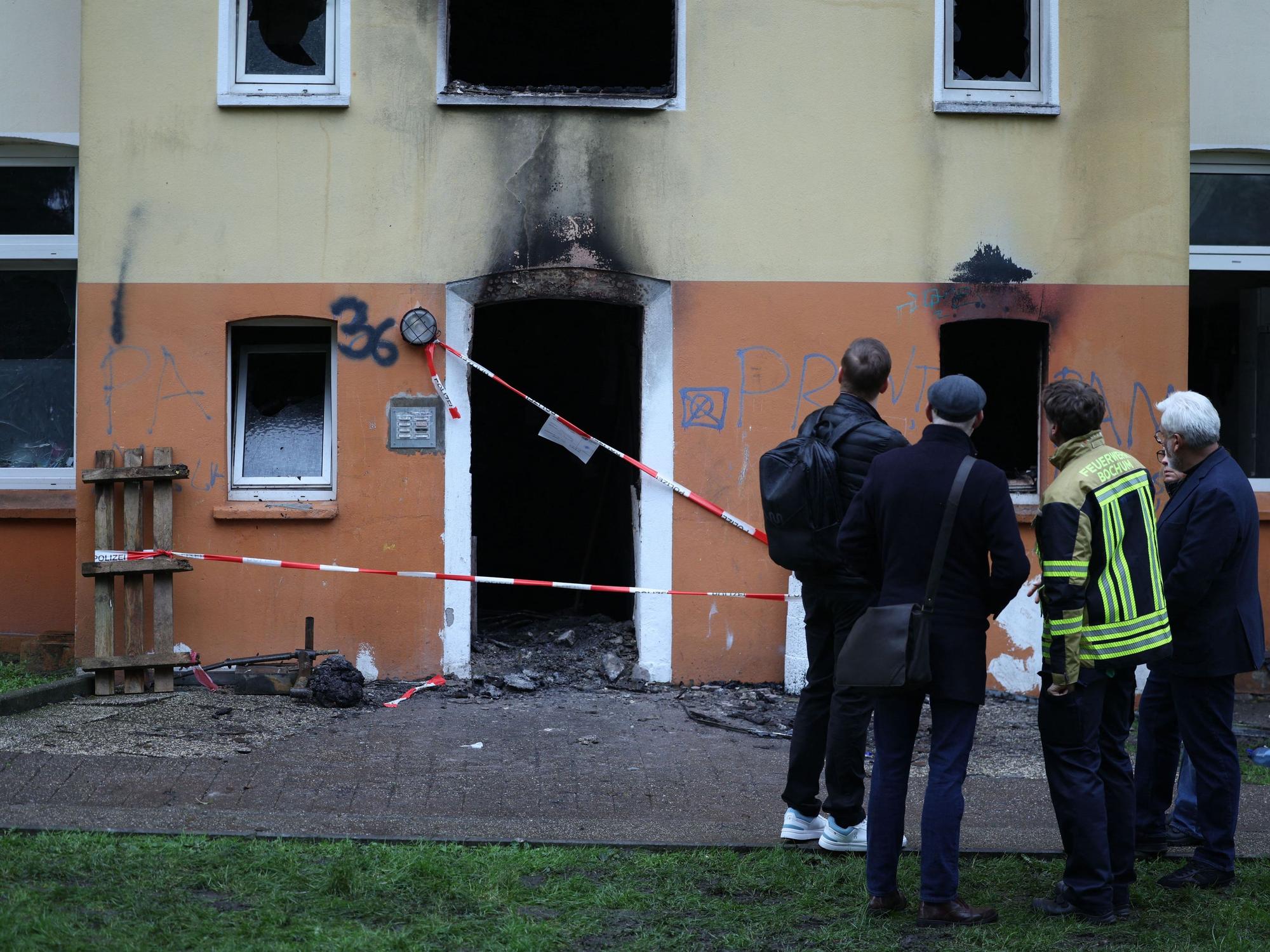 Vier Männer, einer in Feuerwehrjacke, stehe vor dem Mehrfamilienhaus, in dem es gebrannt hat. Der Eingang ist mit Polizei-Flatterband abgesperrt, an der Fassade klebt Ruß.