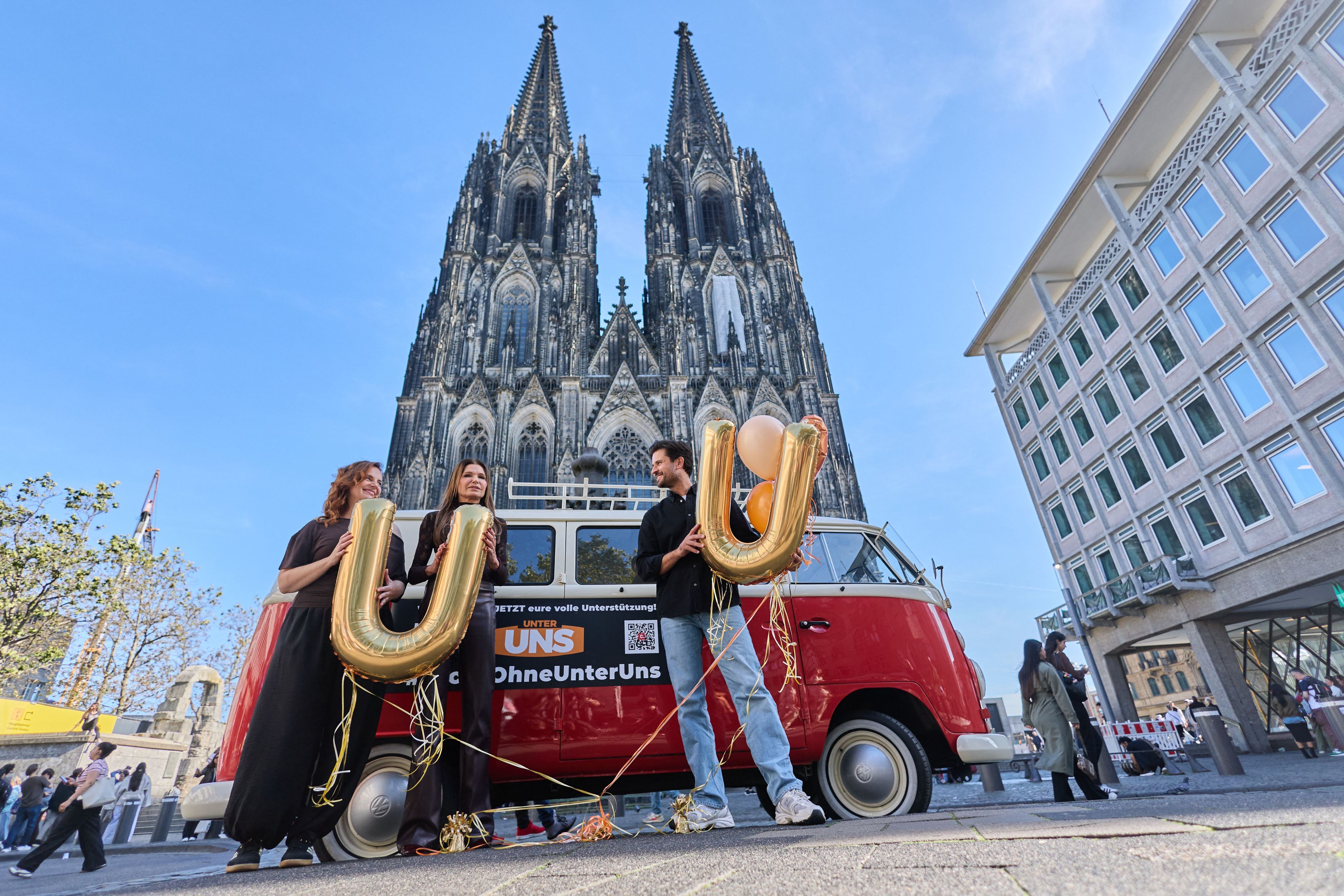 16.10.2026, Nordrhein-Westfalen, Köln: Milos Vukovic (r), Marie Zielcke (l) und Tabea Heynig, Schauspieler der RTL Daily Soap „Unter Uns“, stehen mit einem alten VW Bulli vor dem Dom. Die Schauspieler wollen für eine Fortführung der Serie werben, die RTL nach mehr als drei Jahrzehnten aus dem Programm nehmen wird. Foto: Rolf Vennenbernd/dpa +++ dpa-Bildfunk +++