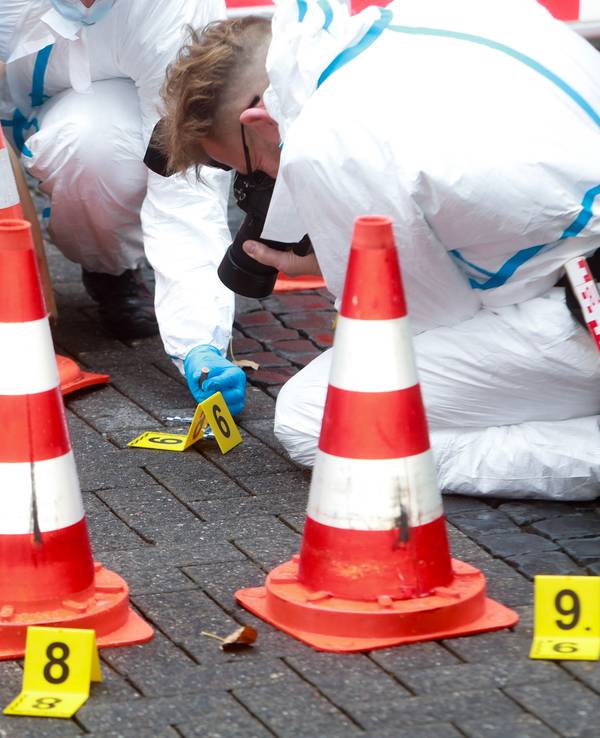 Hütchen und nummerierte Schilder kennzeichen Spuren auf einem Steinboden, ein Beamter der Spurensicherung macht Fotos.