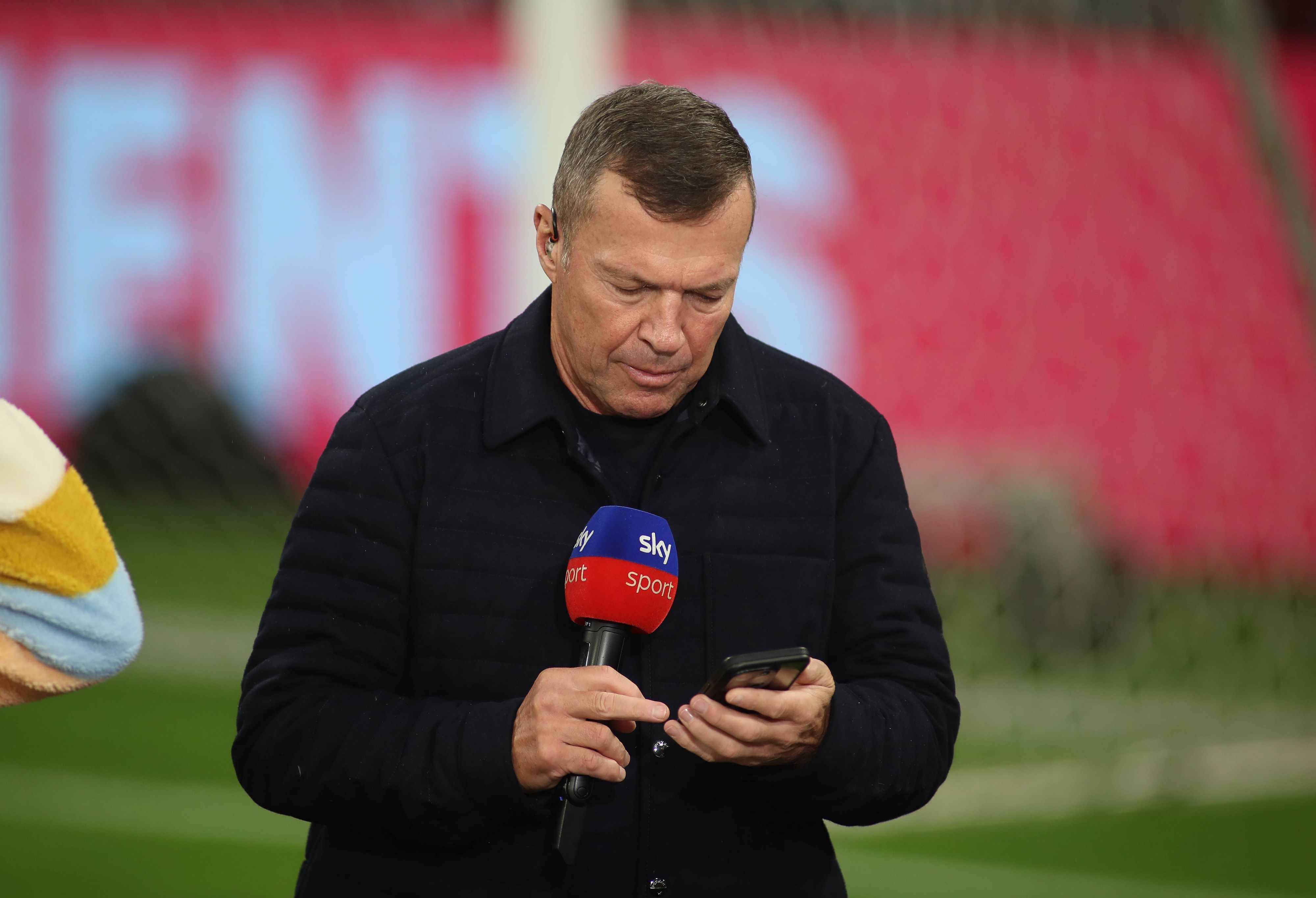 Lothar Matthäus guckt in sein Handy. In der anderen Hand hält er ein Sky-Mikrofon.