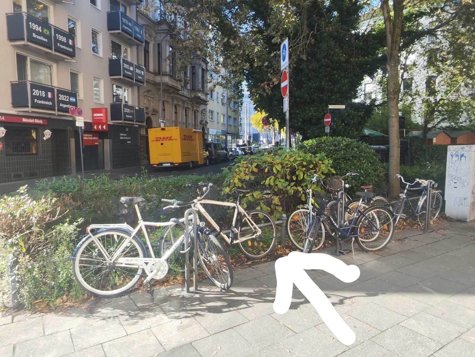 Ein Pfeil zeigt auf eine Stelle zwischen geparkten Fahrrädern.