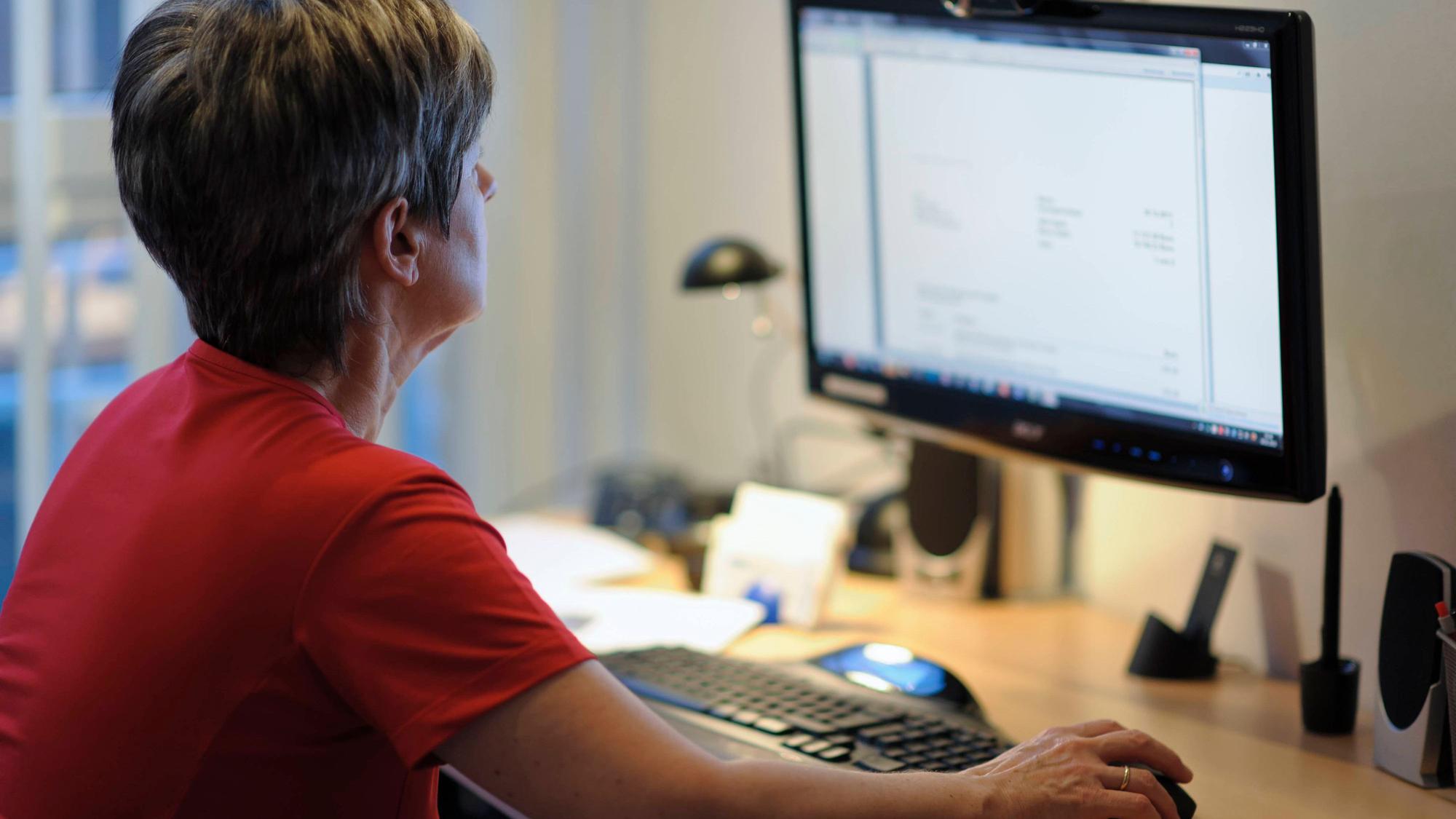 Eine Frau sitzt am Schreibtisch vor einem PC, die rechte Hand liegt auf der Maus.