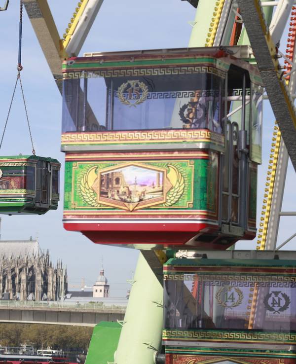 Eine Gondel des Riesenrads wird von einem Kran angehoben, im Hintergrund ist der Kölner Dom zu sehen.