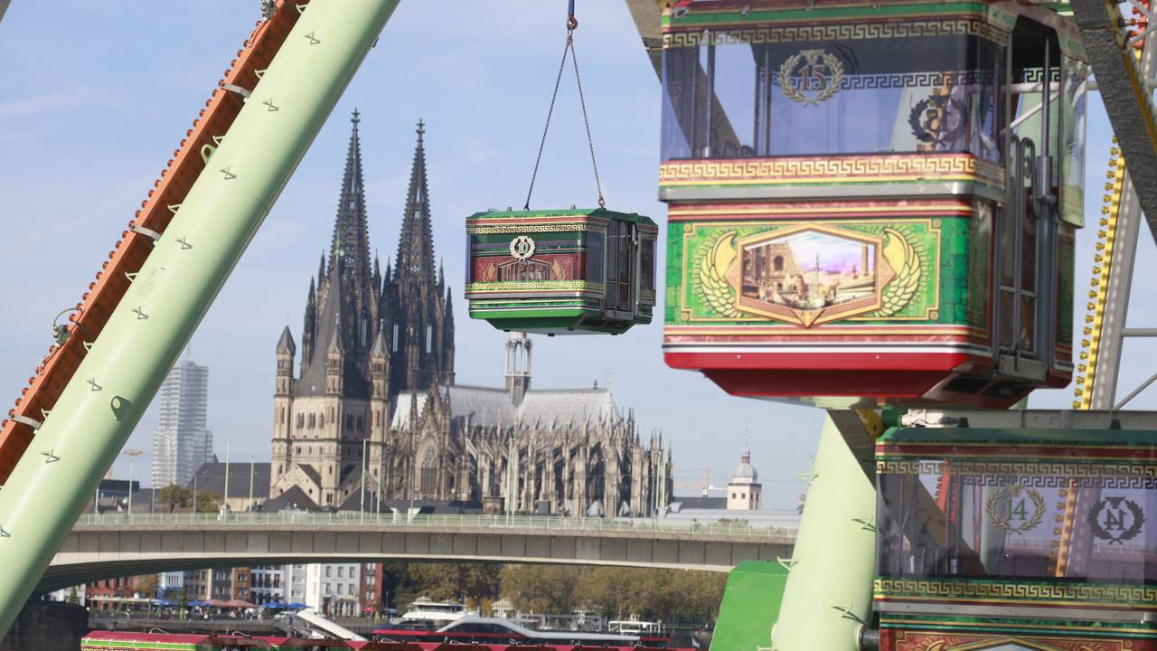 Eine Gondel des Riesenrads wird von einem Kran angehoben, im Hintergrund ist der Kölner Dom zu sehen.