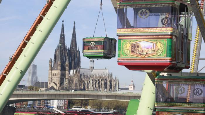 Eine Gondel des Riesenrads wird von einem Kran angehoben, im Hintergrund ist der Kölner Dom zu sehen.