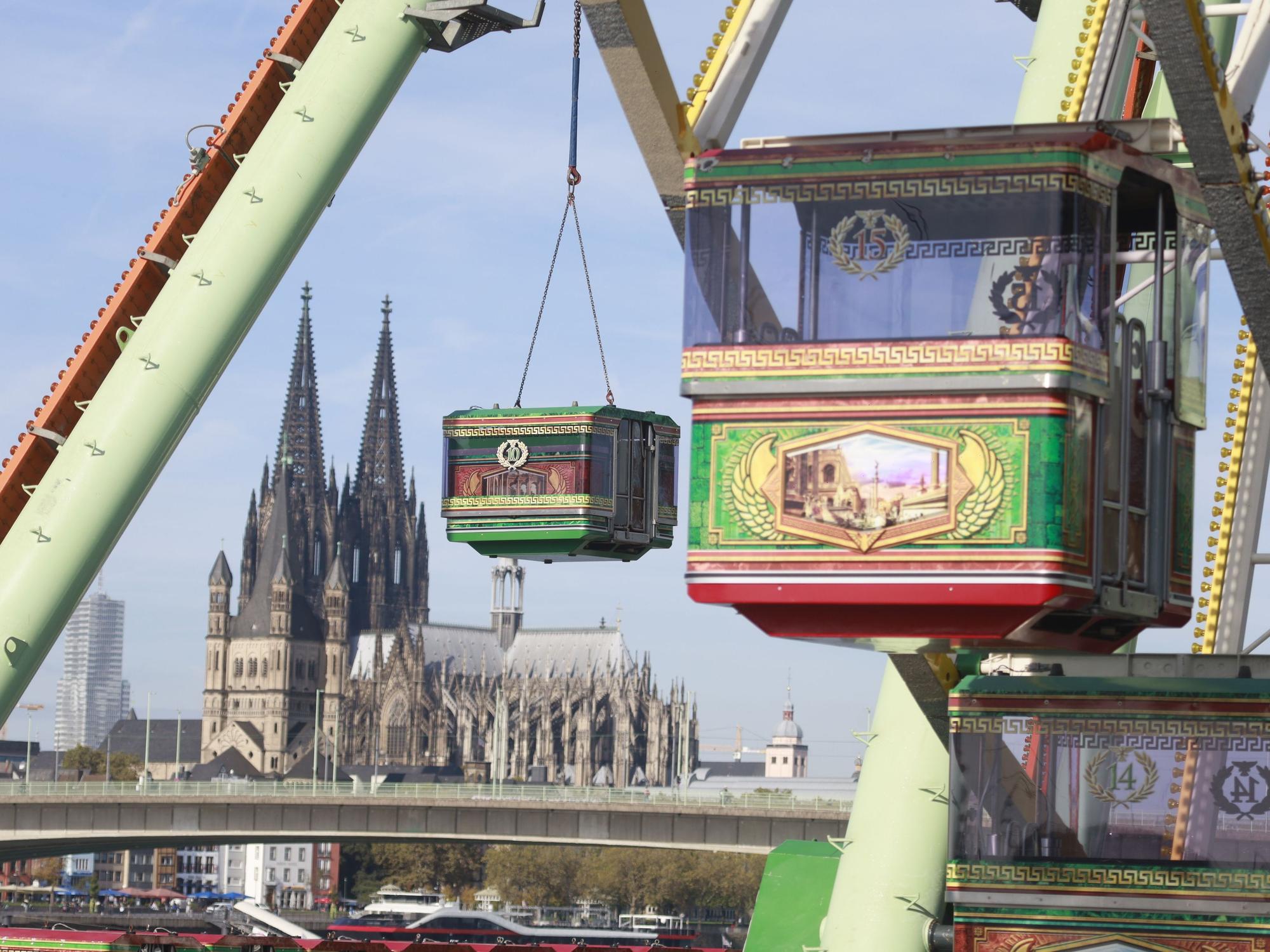 Eine Gondel des Riesenrads wird von einem Kran angehoben, im Hintergrund ist der Kölner Dom zu sehen.