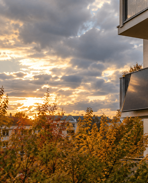 Mehrstöckiges Wohnhaus im Herbst mit zwei schwarzen Solarmodulen außen an der Balkonbrüstung, darüber ein bewölkter Himmel mit goldenen Sonnenstrahlen zwischen den Wolken, im Vordergrund bunte Herbstbäume