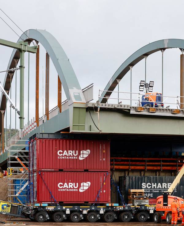 Die neue Stabbogenbrücke an der Landesstraße 193 in Königswinter steht auf Schwerlasttransportern. Bauarbeiter stehen unter der Brückenkonstruktion. Straßen.NRW lässt die neue Brücke über die Bahngleise der Deutschen Bahn einschieben. Die Brücke ist Teil einer wichtigen Verkehrsverbindung zwischen Bonn und Königswinter.