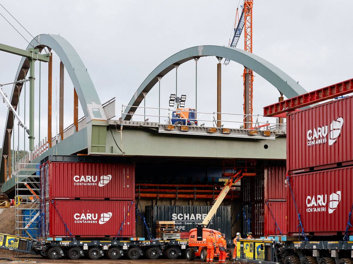 Die neue Stabbogenbrücke an der Landesstraße 193 in Königswinter steht auf Schwerlasttransportern. Bauarbeiter stehen unter der Brückenkonstruktion. Straßen.NRW lässt die neue Brücke über die Bahngleise der Deutschen Bahn einschieben. Die Brücke ist Teil einer wichtigen Verkehrsverbindung zwischen Bonn und Königswinter.