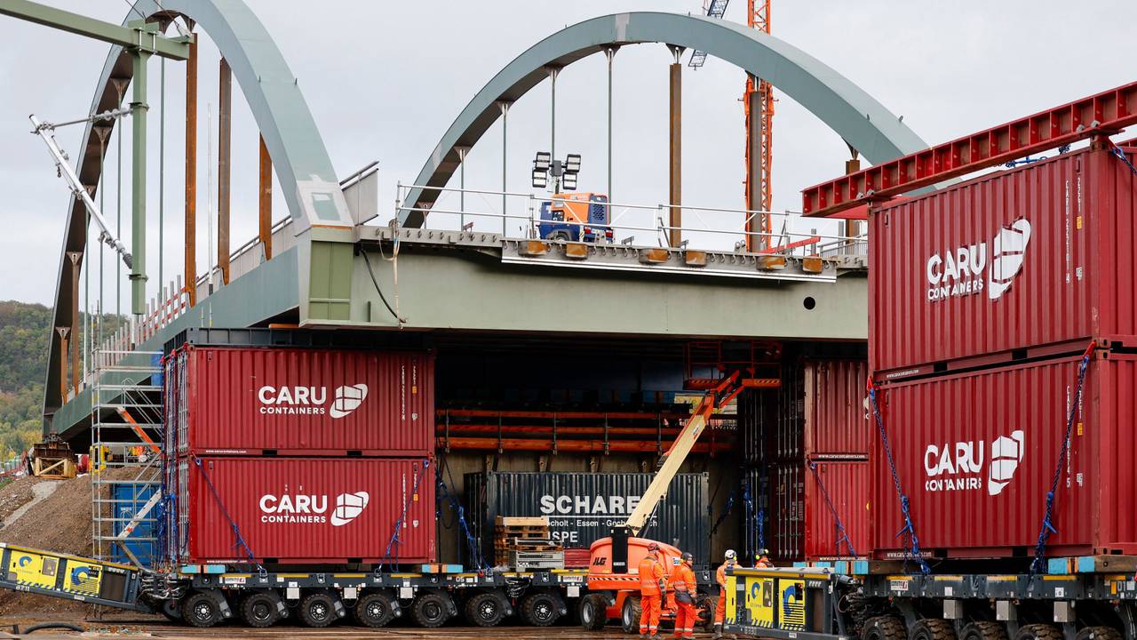 Die neue Stabbogenbrücke an der Landesstraße 193 in Königswinter steht auf Schwerlasttransportern. Bauarbeiter stehen unter der Brückenkonstruktion. Straßen.NRW lässt die neue Brücke über die Bahngleise der Deutschen Bahn einschieben. Die Brücke ist Teil einer wichtigen Verkehrsverbindung zwischen Bonn und Königswinter.