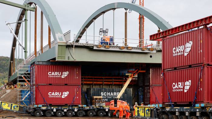 Die neue Stabbogenbrücke an der Landesstraße 193 in Königswinter steht auf Schwerlasttransportern. Bauarbeiter stehen unter der Brückenkonstruktion. Straßen.NRW lässt die neue Brücke über die Bahngleise der Deutschen Bahn einschieben. Die Brücke ist Teil einer wichtigen Verkehrsverbindung zwischen Bonn und Königswinter.