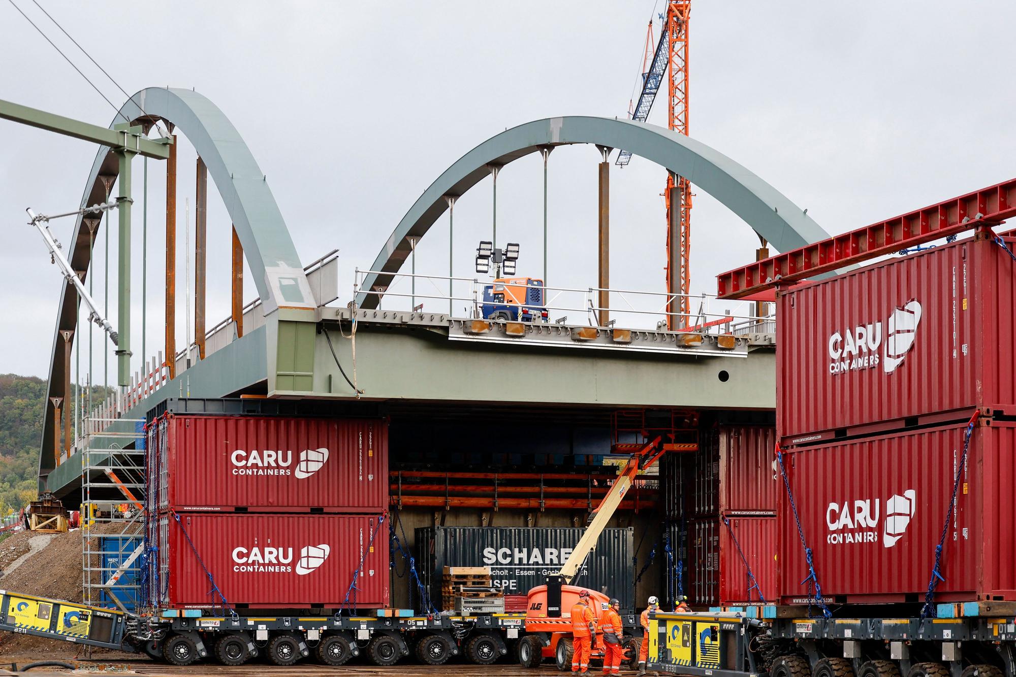 Die neue Stabbogenbrücke an der Landesstraße 193 in Königswinter steht auf Schwerlasttransportern. Bauarbeiter stehen unter der Brückenkonstruktion. Straßen.NRW lässt die neue Brücke über die Bahngleise der Deutschen Bahn einschieben. Die Brücke ist Teil einer wichtigen Verkehrsverbindung zwischen Bonn und Königswinter.
