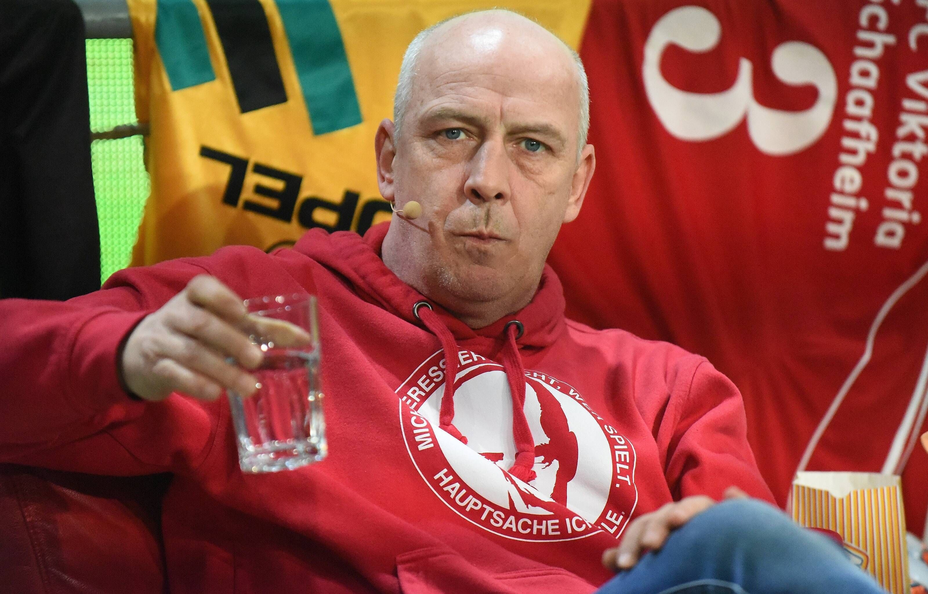 Mario Basler trinkt bei einer Veranstaltung auf der Bühne einen Schluck Wasser.