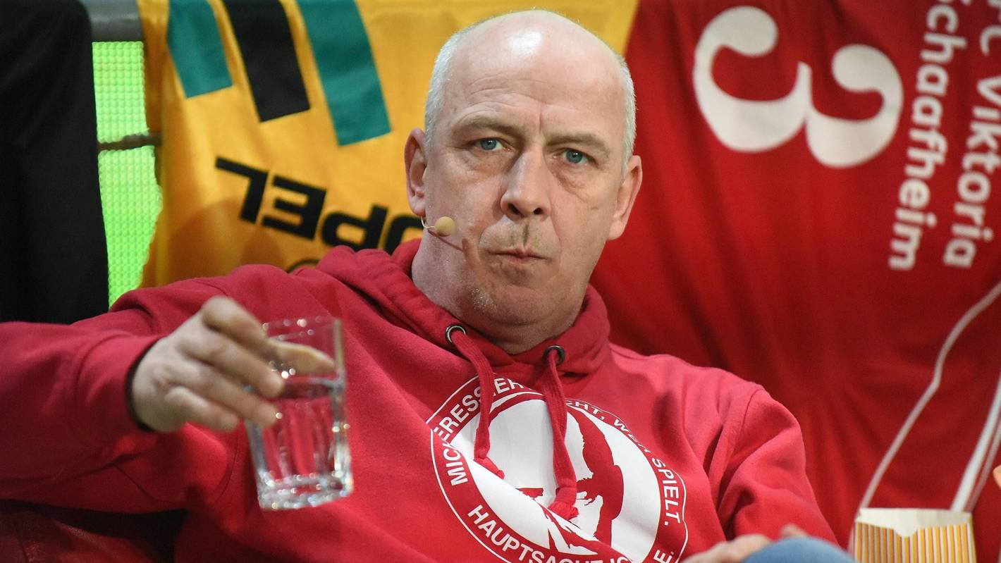 Mario Basler trinkt bei einer Veranstaltung auf der Bühne einen Schluck Wasser.
