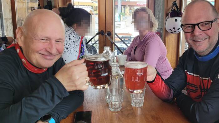 Zwei Männer sitzen an einem Tisch und stoßen mit Biergläsern an, im Hintergrund sitzen zwei Frauen.