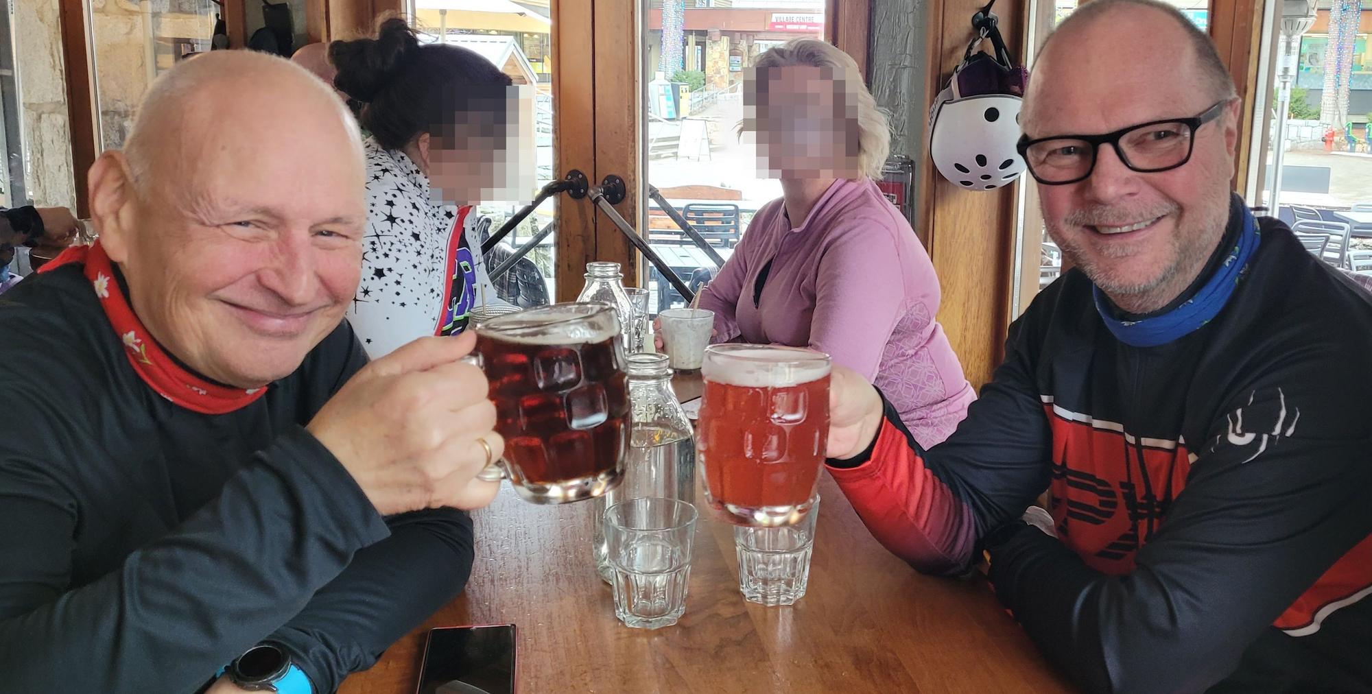 Zwei Männer sitzen an einem Tisch und stoßen mit Biergläsern an, im Hintergrund sitzen zwei Frauen.