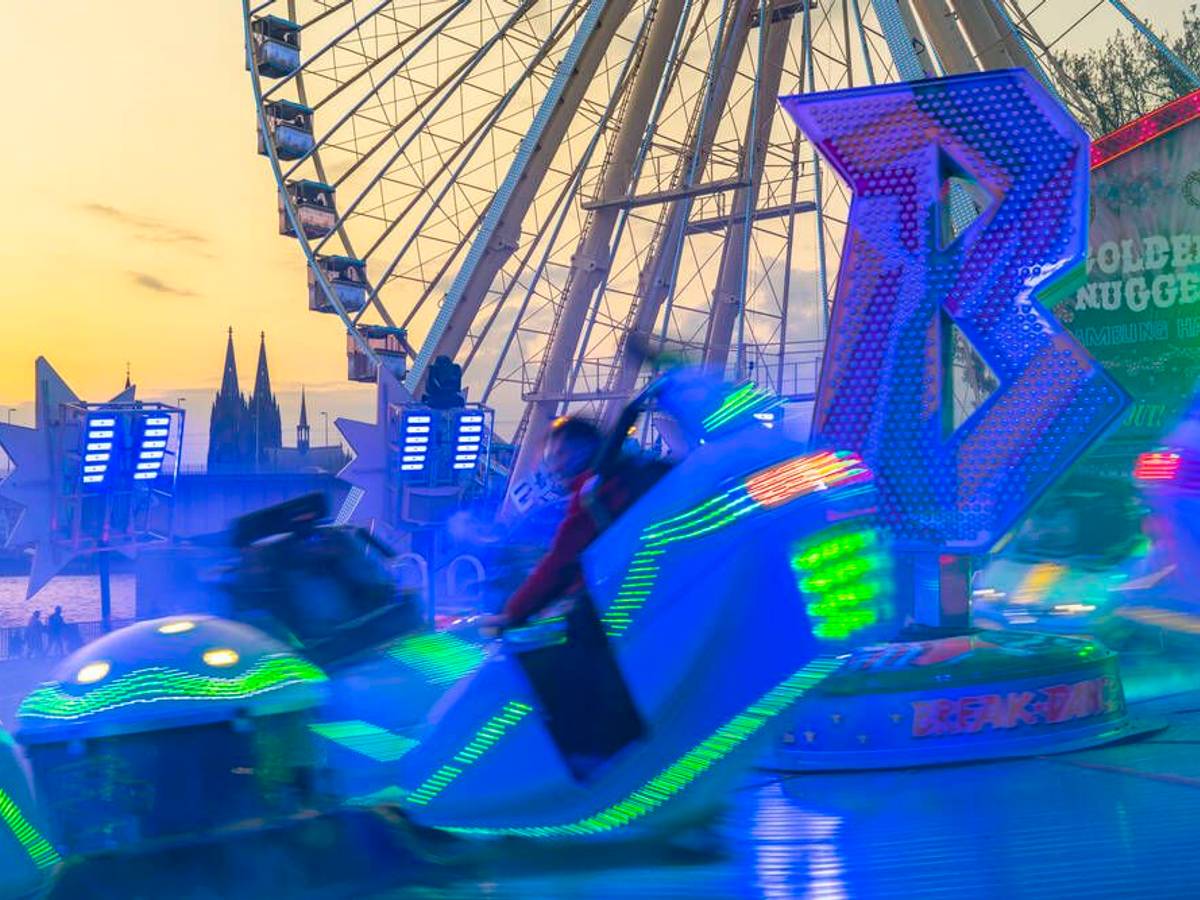 Ein leuchtendes Fahrgeschäft vor einem Riesenrad und dem Kölner Dom.