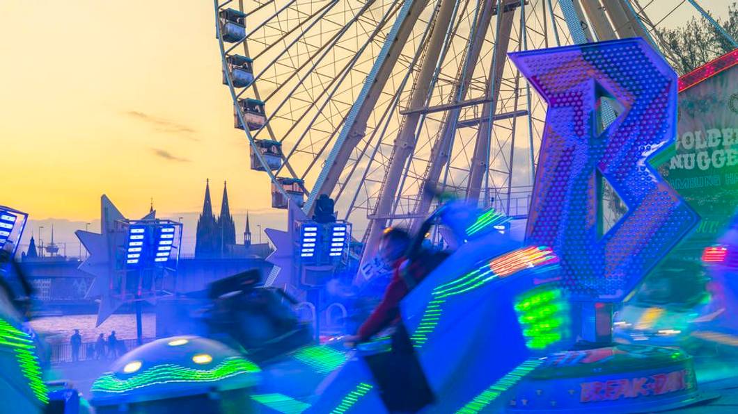 Ein leuchtendes Fahrgeschäft vor einem Riesenrad und dem Kölner Dom.