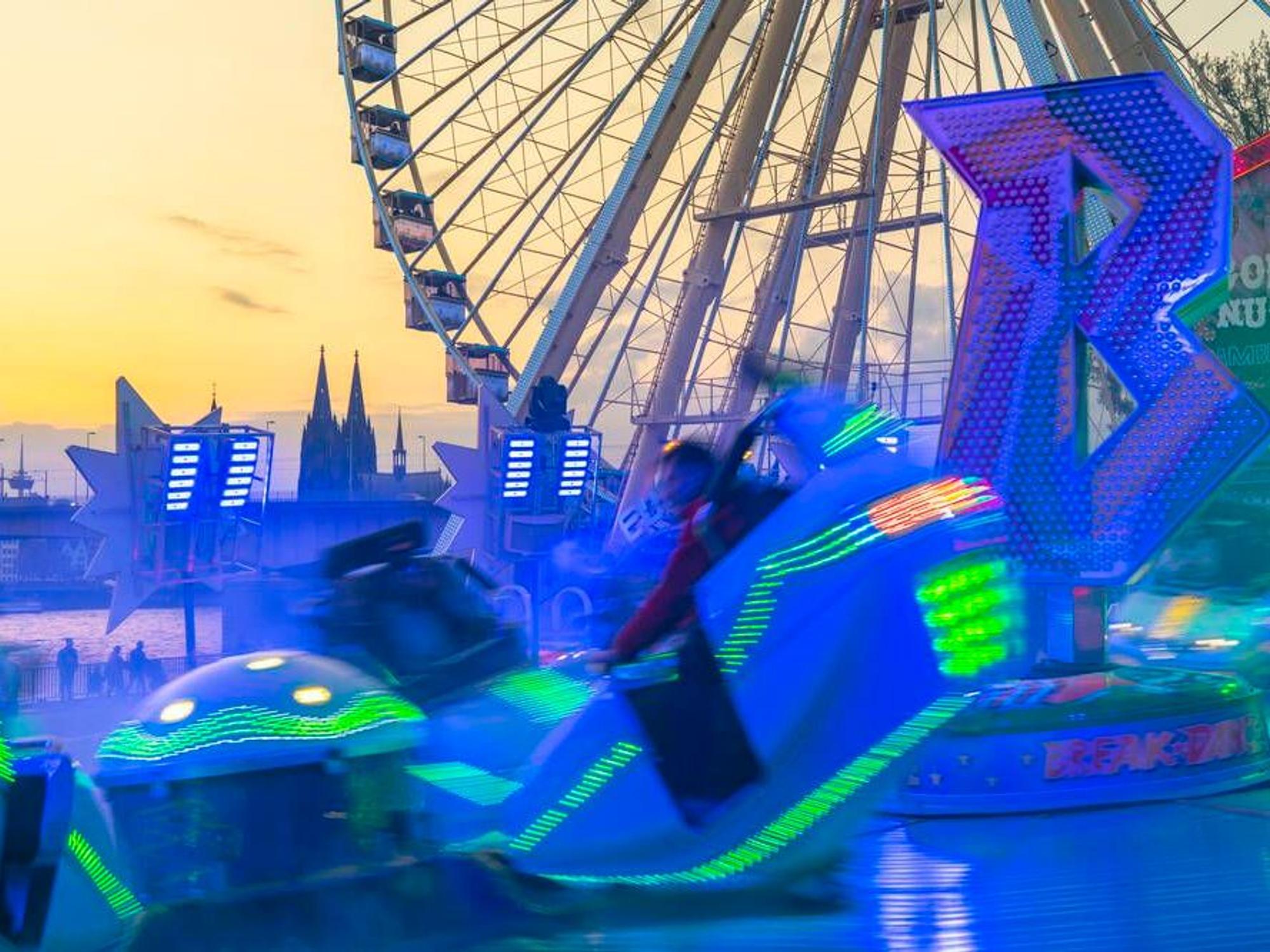 Ein leuchtendes Fahrgeschäft vor einem Riesenrad und dem Kölner Dom.