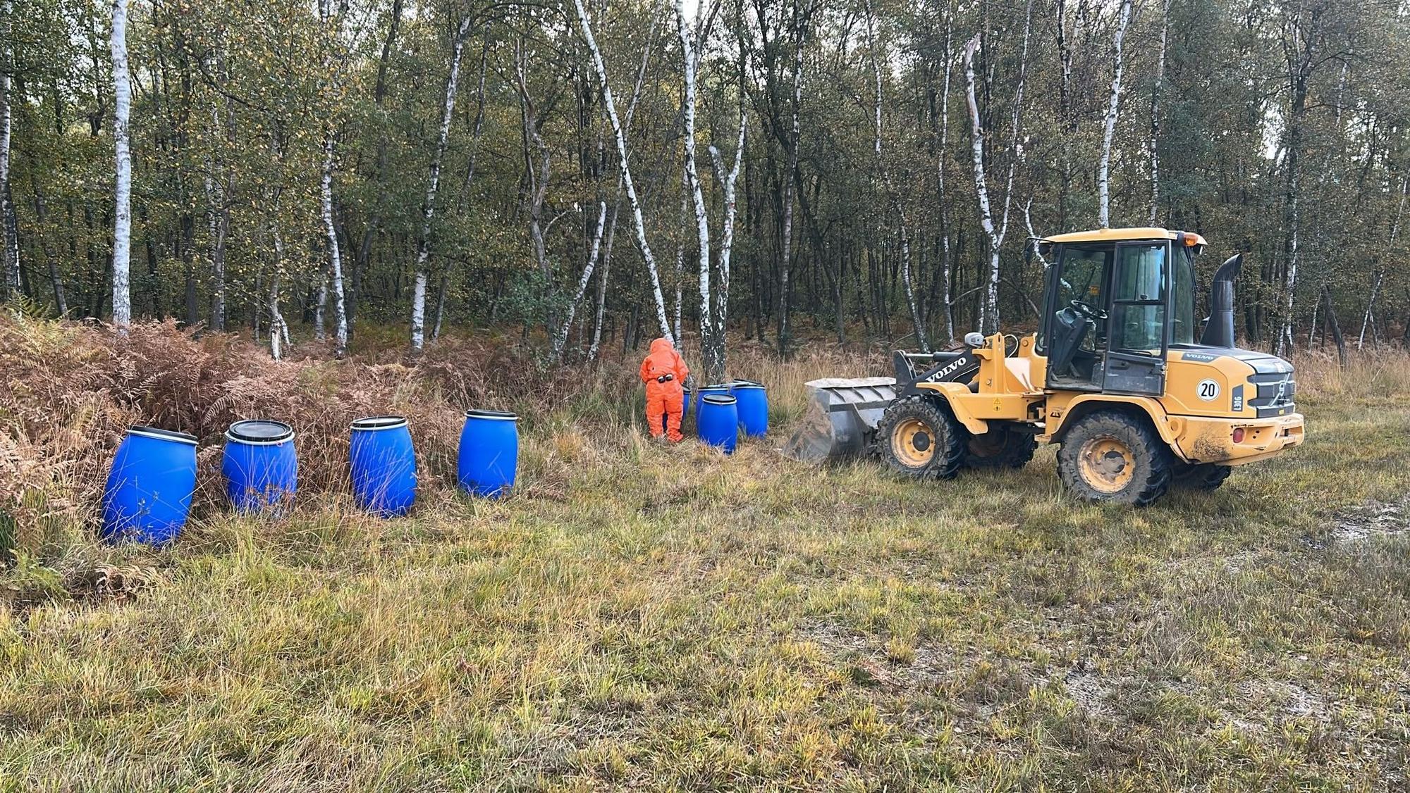 Mehrere blaue Fässer stehen im Naturschutzgebiet, ein Mann im Schutzanzug steht daneben.
